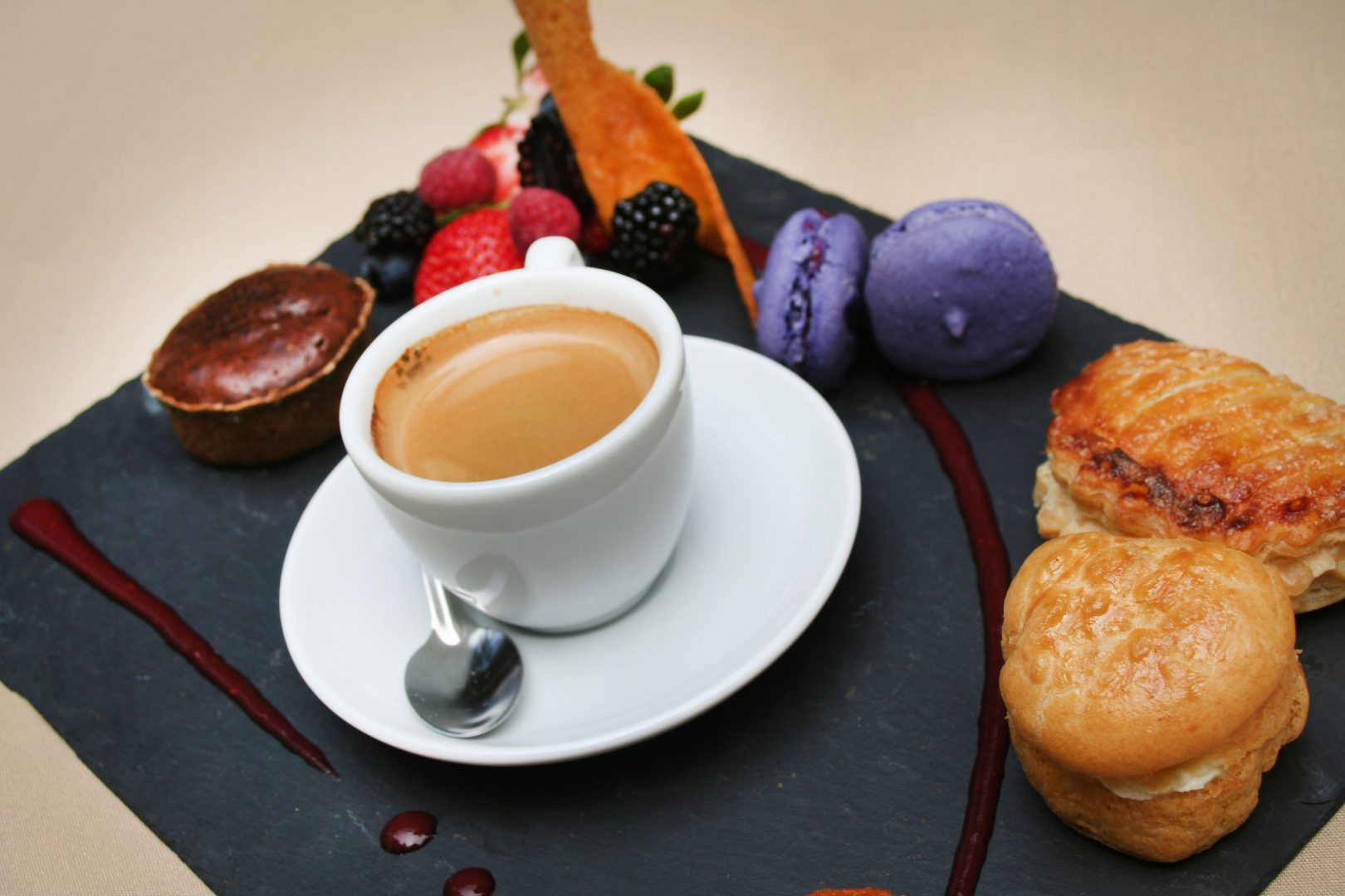 Espresso cup with assorted pastries, berries, and macarons arranged on a slate platter with berry coulis.