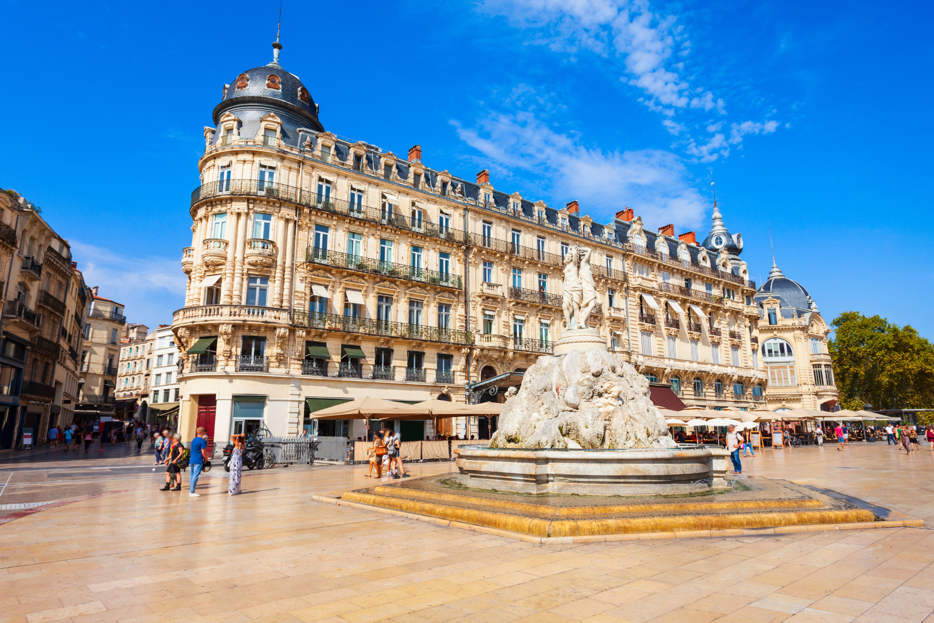 A detailed ornate building overlooks a stone fountain with statues in a sunny plaza with people walking.