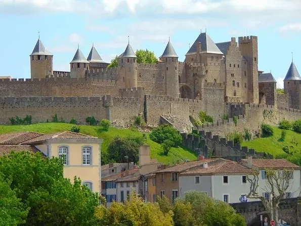 A medieval stone castle with conical towers sits atop a grassy hill, overlooking a town with terracotta roofs and yellow buildings.
