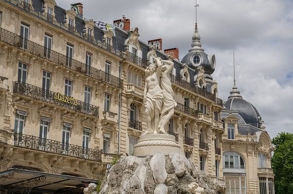 A stone sculpture of two women stands in front of a grand building with ornate balconies and a domed roof. "La Gazette" is visible on the building.