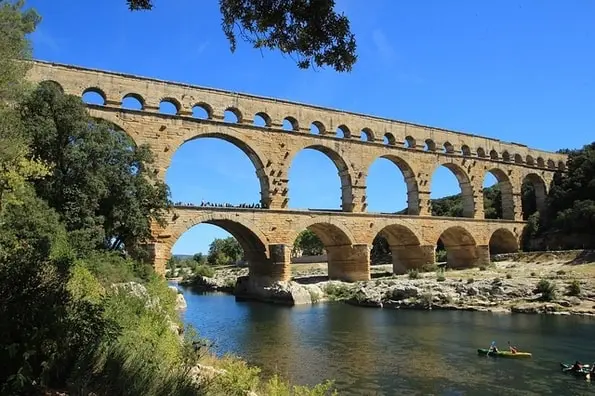 The Pont du Gard, a three-tiered Roman aqueduct made of stone, spans a clear blue river under a bright sky. Kayakers are visible on the water.