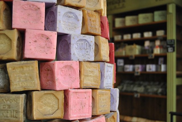 A stack of colorful, cube-shaped soaps with embossed text, including 'Savon de Marseille' and '300'. Shelves of similar soaps are visible in the blurred background.