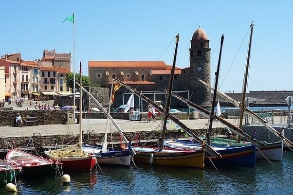 Colorful sailboats docked in a harbor with a stone building and clock tower in the background.