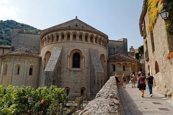 Stone church with rounded apse and arched windows, people walk along a path beside it.