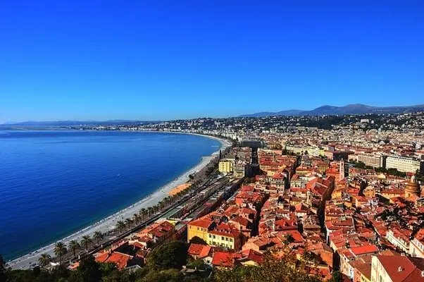 Panoramic view of Nice, France, with the blue Mediterranean Sea, a pebble beach, palm trees, and a dense city skyline under a clear blue sky.