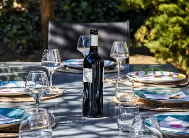 A bottle of red wine stands on a table set for a meal outdoors, with plates and glasses.
