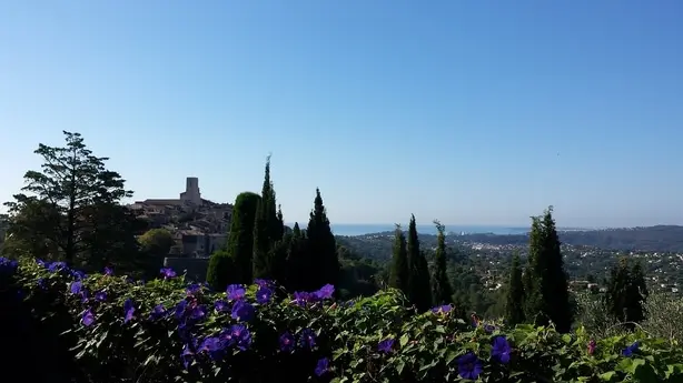 A village with a prominent tower sits on a hill, overlooking a coastal town and the sea. Purple flowers bloom in the foreground.