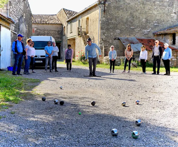 Players in a village courtyard play pétanque, with metal balls scattered on the gravel and a man in a blue shirt throwing.