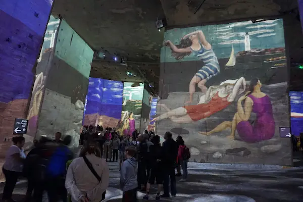 A large projection of vintage beach scenes with women in swimsuits covers the walls of an indoor space. A crowd of people observe the display.