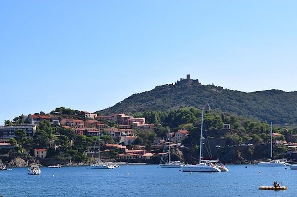 Coastal town with red-roofed buildings nestled on a hillside, crowned by a distant castle ruin. Sailboats dot the blue water.