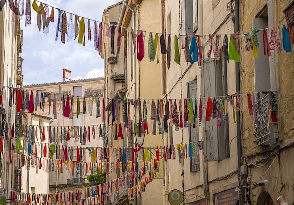 Colorful fabric strips hang on clotheslines strung across a narrow street between buildings.