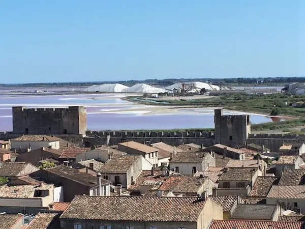 A medieval walled town with terracotta roofs overlooks pink salt flats and large white salt piles under a clear blue sky.
