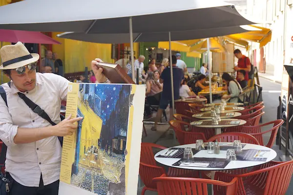 Man in hat points to Van Gogh's "Cafe Terrace at Night" painting displayed outdoors at a cafe with patrons dining.