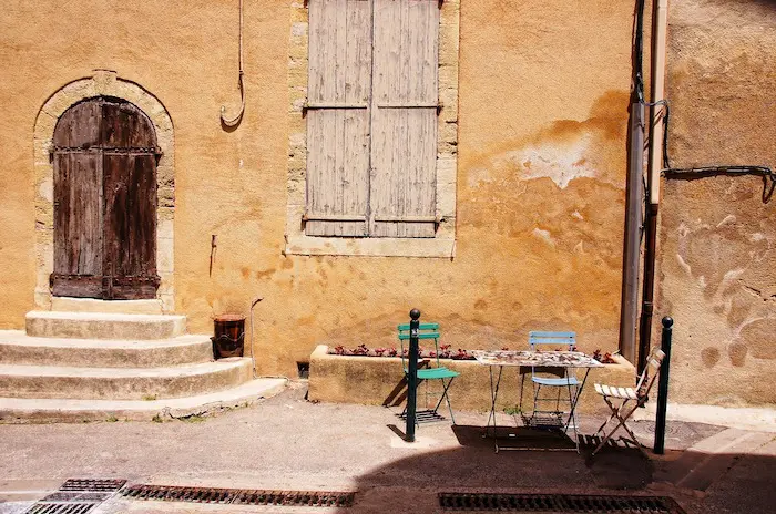 A rustic wooden door with arched stone frame and three steps leads to a building with weathered shutters. A small cafe table with chairs sits outside.