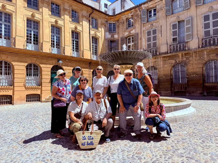 A diverse group of ten people poses for a photo in a courtyard with a fountain. A basket reads La vie est belle.