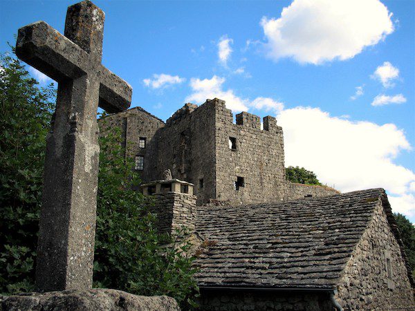 A weathered stone cross stands in the foreground, with a medieval stone village and castle ruins in the background under a blue sky with clouds.