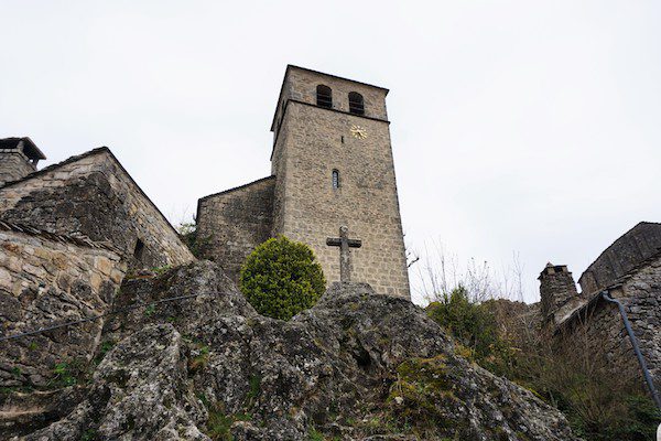 Stone church tower with a clock face, a cross, and surrounding stone buildings on a rocky hillside.