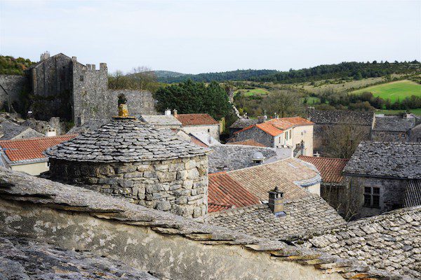 Stone buildings with slate roofs and a castle in the background, overlooking a green, rolling landscape.