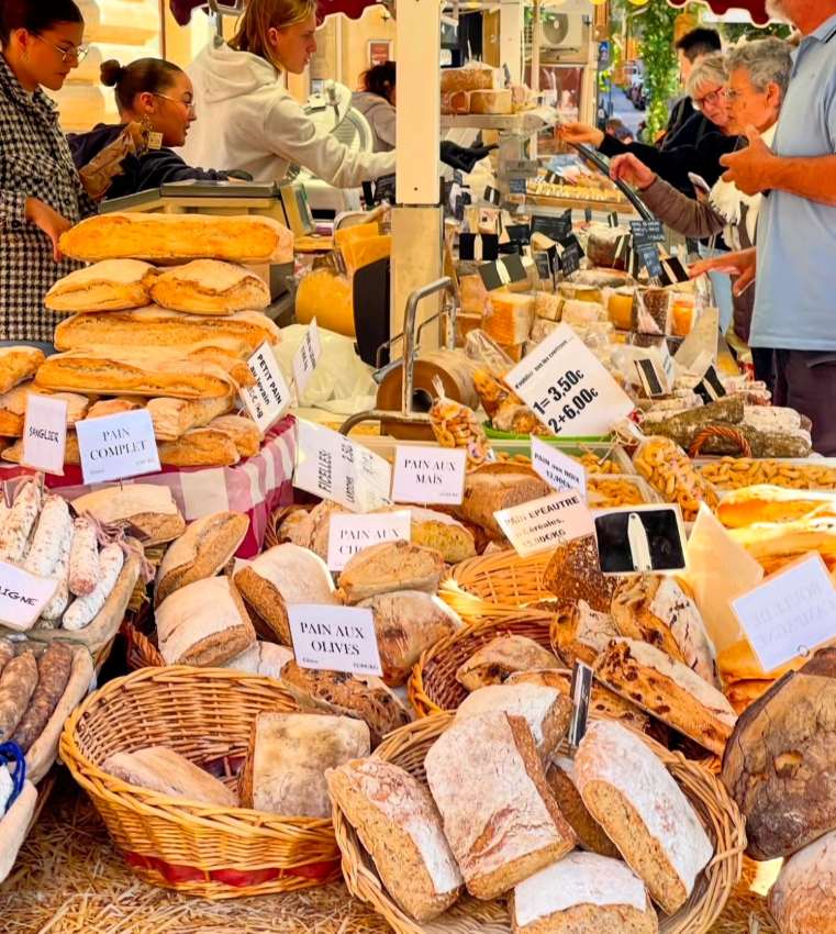 A vibrant outdoor market stall overflows with freshly baked breads, baguettes, and loaves. Various types of bread are displayed in baskets and on tables. Shoppers are seen selecting their purchases.
