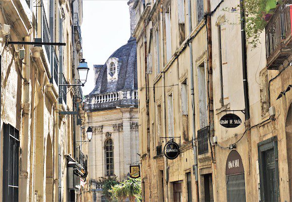 Narrow European street with old buildings, a domed roof structure, and signs for Guinness and Grain de Soleil.