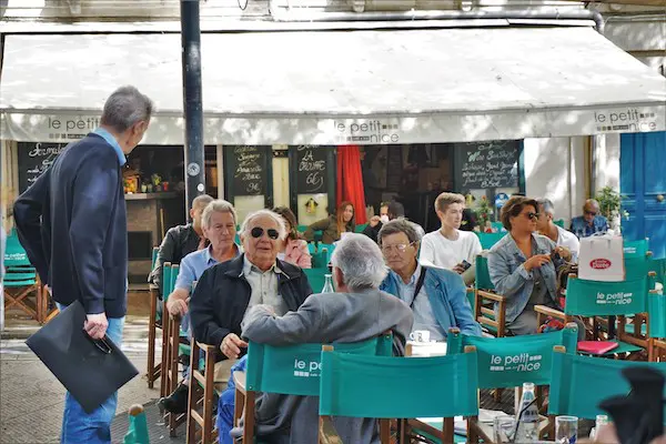 Several people sit at tables outside a cafe. A man in a blue jacket stands holding a black folder.