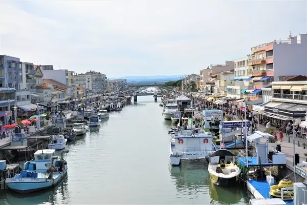 Boats line a canal in a European town with shops and restaurants on both sides. A bridge spans the canal in the distance.