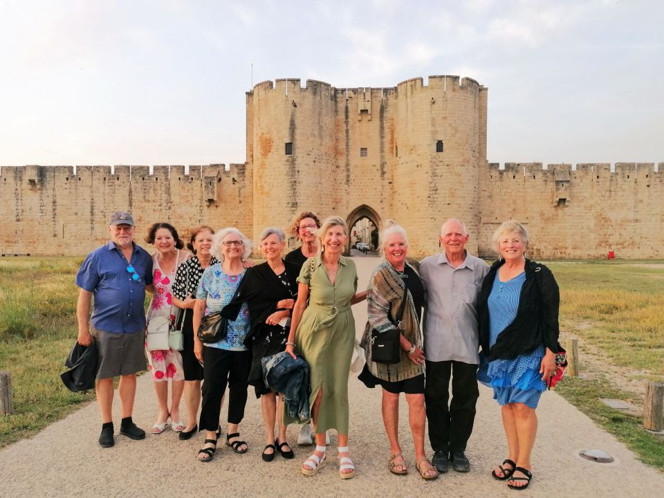 A group of ten smiling adults poses for a photo in front of a large stone fortified gate and wall of Aigues Mortes