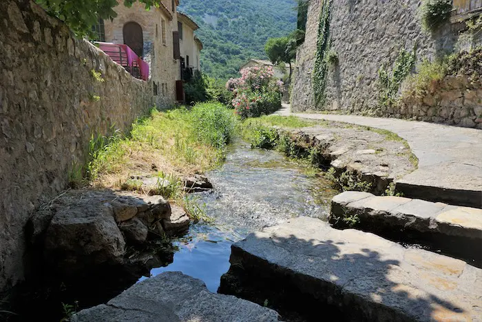 A clear stream flows through a village setting with stone walls, a curved pathway, and lush greenery leading to distant hills.