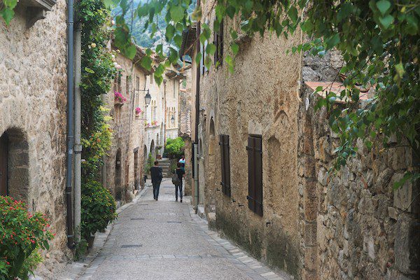 A narrow cobblestone street winds through a historic village with stone buildings adorned with flowers and greenery.