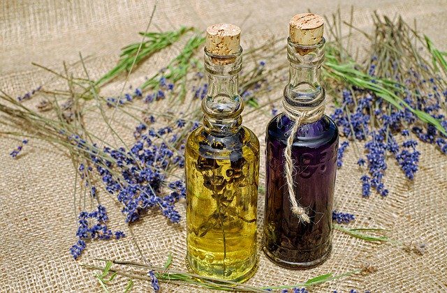 Two glass bottles with cork stoppers, one filled with yellow liquid and herbs, the other with purple liquid. Surrounded by lavender and rosemary sprigs on burlap.