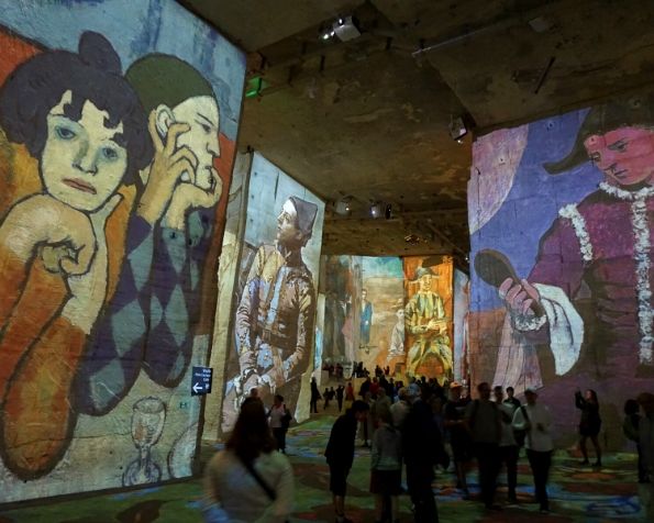 People walk through a large space where paintings are projected onto walls, including portraits of figures in colorful attire.