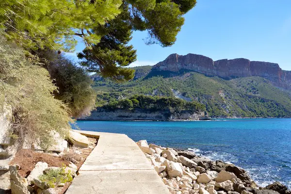 A paved path curves along a rocky coastline beside bright blue water, with green trees and a large, flat-topped mountain in the background.