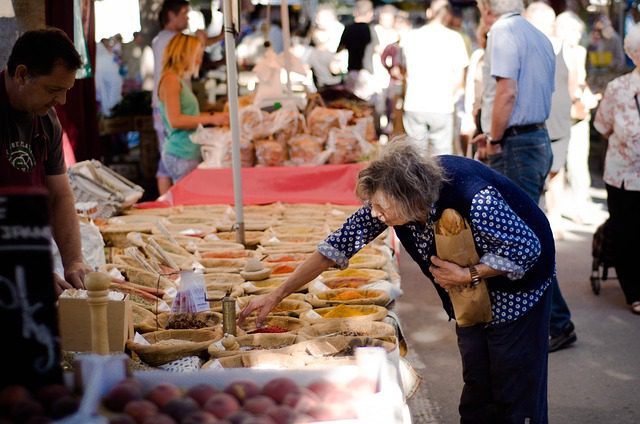 An elderly woman with gray hair bends over a market stall, reaching for spices. She holds a paper bag with a baguette.