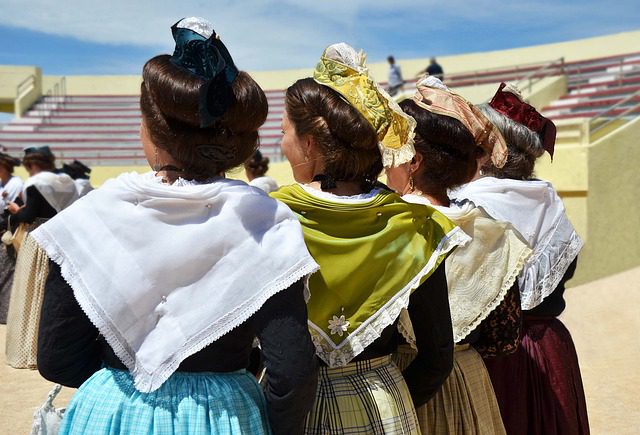 Women in traditional dress with elaborate hairstyles and colorful headscarves stand in front of stadium seating.