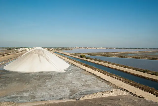 A vast salt pan with a large white mountain of salt in the foreground, surrounded by water channels and ponds under a clear blue sky.