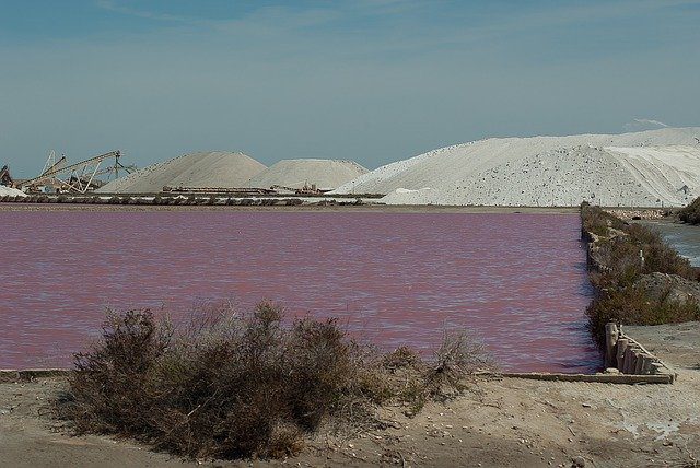 Pink salt pond with large white salt piles and industrial equipment in the background.