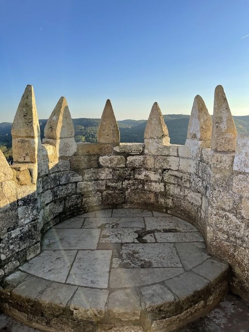 Stone crenellations of a castle tower overlook a green, rolling landscape under a clear blue sky.
