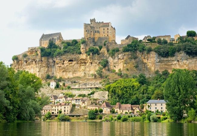 A medieval castle and village perch atop a sheer cliff overlooking a calm river.