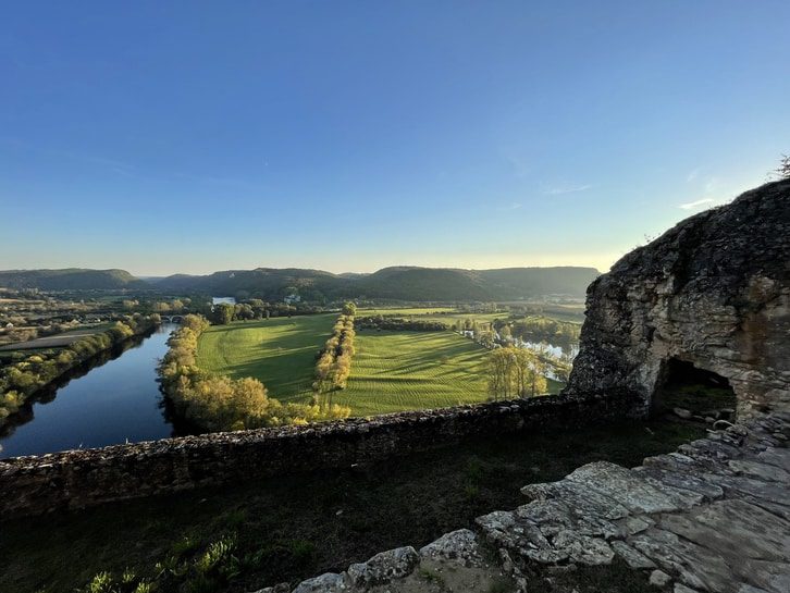View from ancient stone ruins overlooking a wide river, green fields, and rolling hills under a clear blue sky.