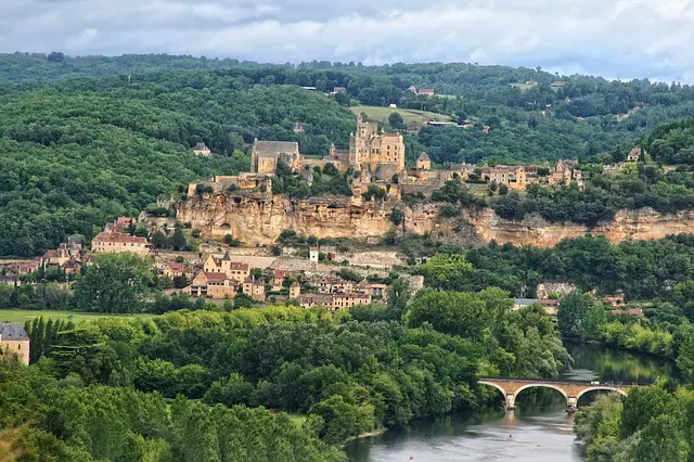 A medieval village and castle perched on a cliffside above a river, surrounded by lush green forests.