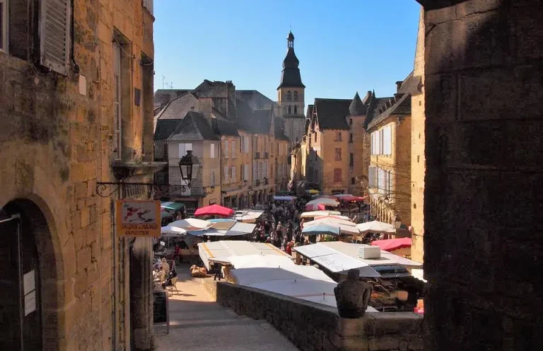 A bustling outdoor market fills a narrow street lined with historic buildings. Stalls with colorful awnings display goods, and a crowd of people browses.