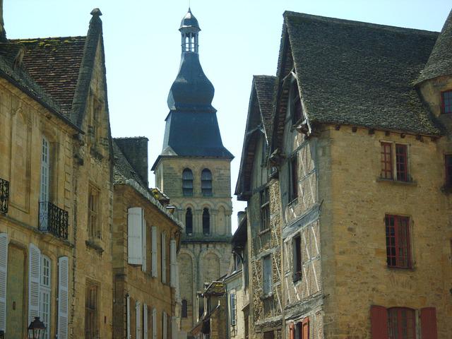 Stone buildings with pitched roofs line a street, leading to a tall bell tower with a dark, domed roof.