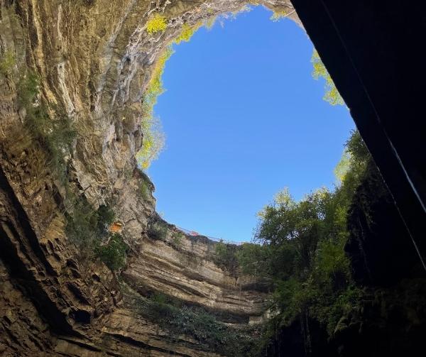 Looking up from inside a cave opening to a bright blue sky, with layered rock walls and green foliage.