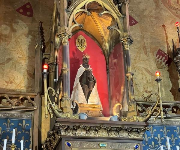A dark statue of a crowned Madonna holding a child, in a golden, ornate shrine with red background and flickering red candles.