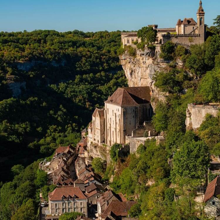 A medieval village clings to a steep cliff face, with a large church and castle overlooking the forested valley.