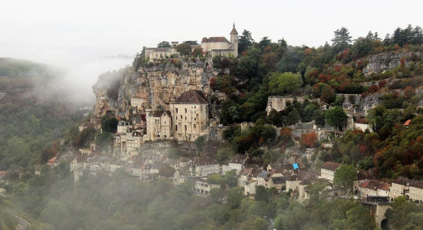 Medieval village clinging to a steep cliff face, with buildings stacked vertically amidst trees and fog.