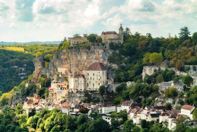 A medieval village clings to a steep cliff face, with buildings and a castle nestled among trees under a cloudy sky.