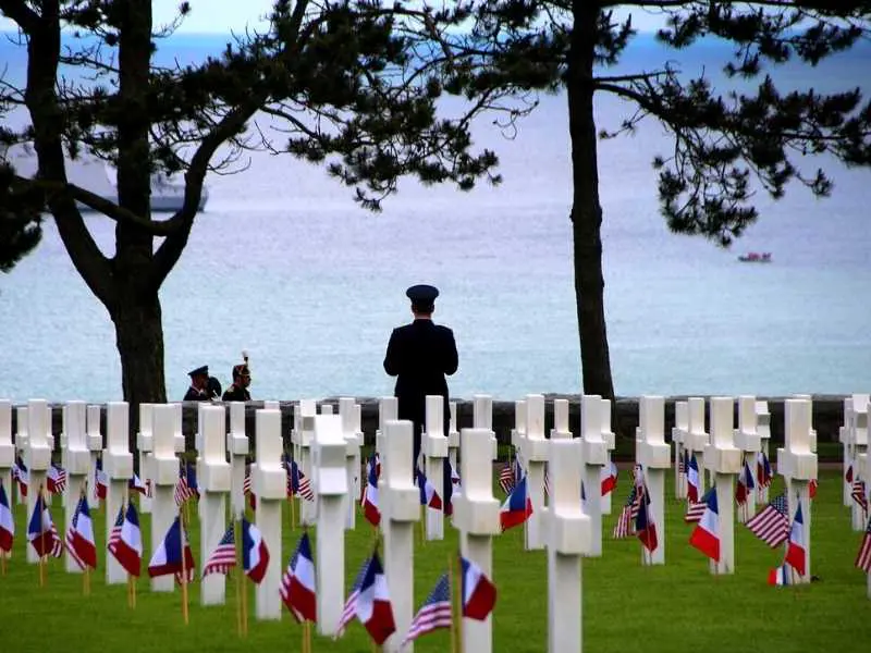 Rows of white crosses with American and French flags stand in a grassy field overlooking the ocean. Two figures in uniform stand behind the crosses.