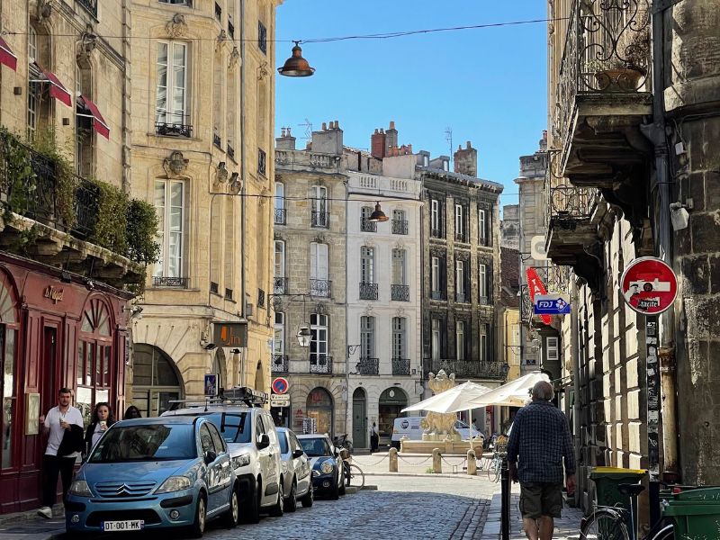 Cobblestone street lined with ornate buildings, cars parked along the side, and a man walking away from the camera.