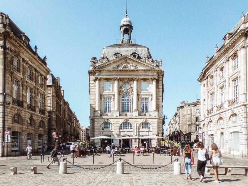 Grand stone buildings flank a cobblestone plaza with people walking and sitting at outdoor cafes.